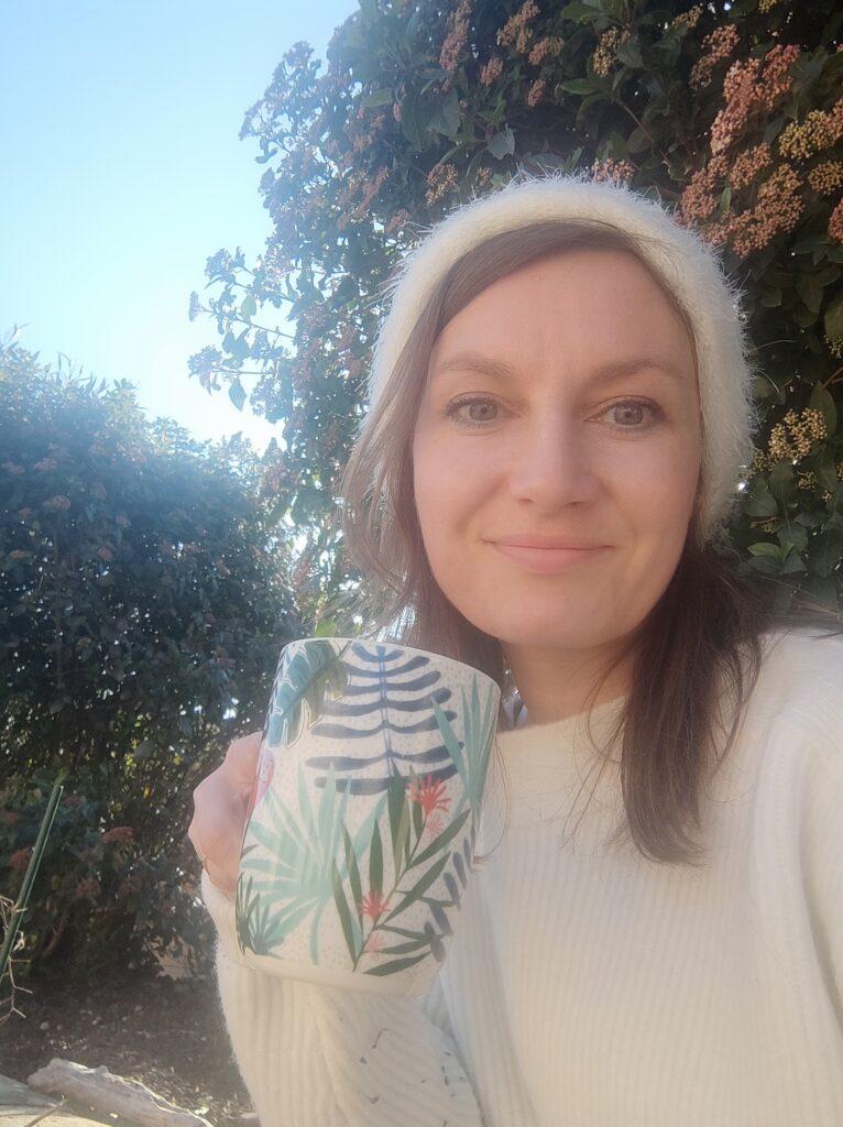 Émilie Lylusio buvant un café, souriante un bonnet en extérieur jardin